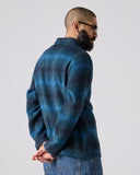 Model wearing peacock blue check, buttoned Pennine wool overshirt. Showing back and arm and paired with jeans.