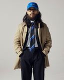Male model with a beard wearing a blue Uskees baseball cap, glasses, beige car coat, midnight blue Uskees pants, and striped tie.