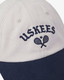 Close-up front view of cream and navy blue Uskees cap with 'USKEES' and tennis racquet motif embroidered in navy blue.
