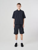 Full-length front view of a male model wearing an Uskees black short-sleeve button front trek shirt paired with matching Uskees technical shorts.