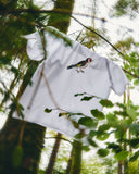 An Uskees white short-sleeve  men's T-shirt hanging outdoors among tree trunks and green leaves, featuring a realistic bird graphic printed on the chest.
