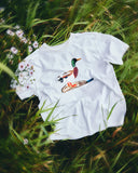 An Uskees white short-sleeve t-shirt for men featuring a printed illustration of a mallard duck laid flat on a bed of green grass with small white flowers. 