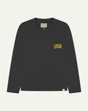 Full flat view of faded black organic cotton signature long sleeve T-shirt for men, showing the varsity inspired 'USK' logo in yellow on the chest pocket, and the Uskees branding labels on the neck and hem.