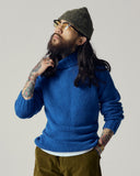 Male model wearing an Uskees ultra blue lambswool sweater and green beanie with tattoos on arms, standing against a plain background.