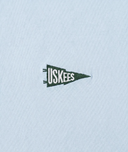 A close-up of a cyan t-shirt featuring a small embroidered dark green and white USKEES pennant logo.