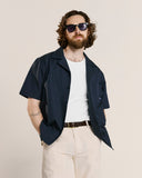 Model wearing an Uskees navy short-sleeve shirt, white t-shirt, beige pants, and sunglasses on a beige background.